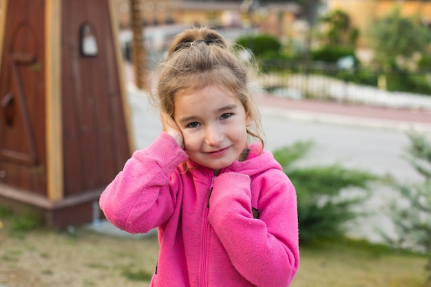 A little girl in a pink hoodie with a sad and tearful face is holding her ear Ear pain otitis media swelling of the cheek gums toothache children's surgery otolaryngology Children's medicine