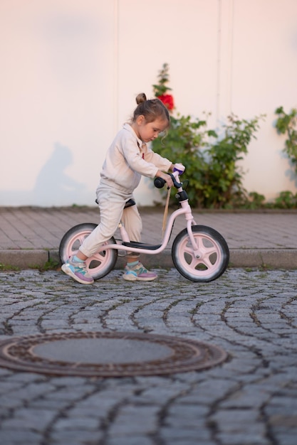 A little girl in a pink suit rides a pink balance bike in the rays of the sunset Child on kids bike