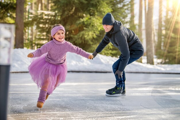 Photo a little girl in a pink sweater and a fluffy skirt on a sunny winter day rides with her dad on an open skating rink in the park