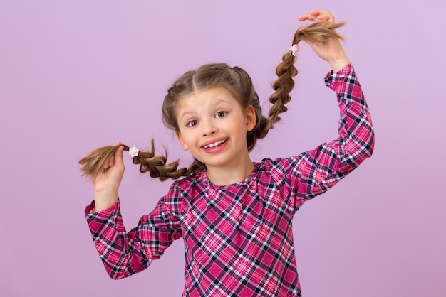 A little girl in a plaid dress holds her braids and makes a face