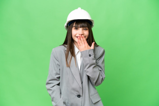 Little girl playing as a architect with helmet and holding blueprints over isolated background happy and smiling covering mouth with hand