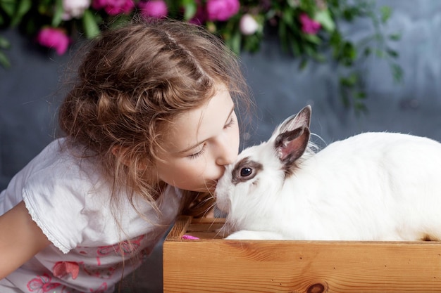 Little girl playing with real rabbit Child and white bunny on Easter on flower background Kid kiss pet Fun and friendship for animals and children