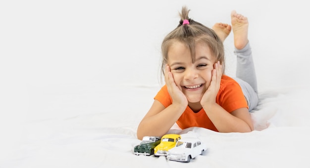 Little girl playing with white toy car. Sitting on the white bedsheet. Smiling.