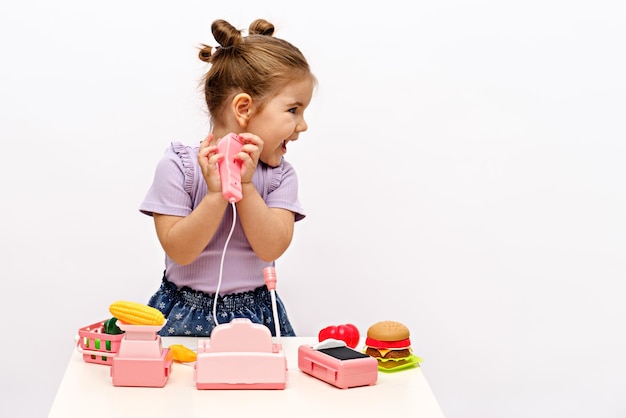 Little girl plays shop with toy cash register scales and plastic fruits and vegetables on white table at home looks away and smiles copy space play store