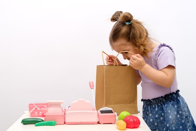 Little girl plays in store looks into bag of groceries toy cash register scales toy vegetables and fruits on table online ordering and delivery of food