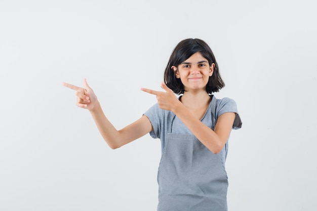 Little girl pointing to the left side in t-shirt, apron and looking merry. front view.