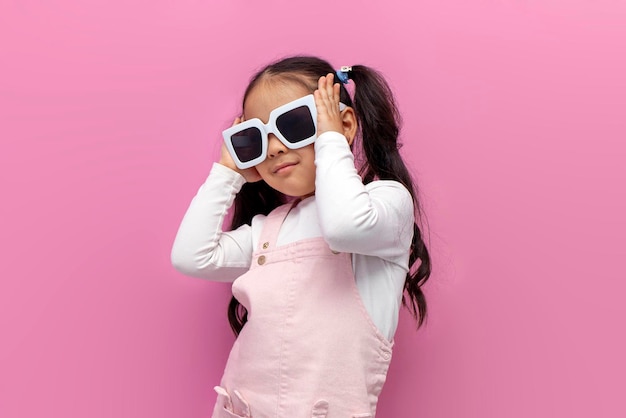 Little girl preschool child in white sunglasses and a pink sundress on a pink isolated background