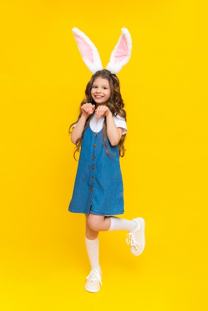 A little girl in a rabbit costume A child with long ears on his head celebrates spring Easter Children's holiday A little girl in full height on a yellow isolated background