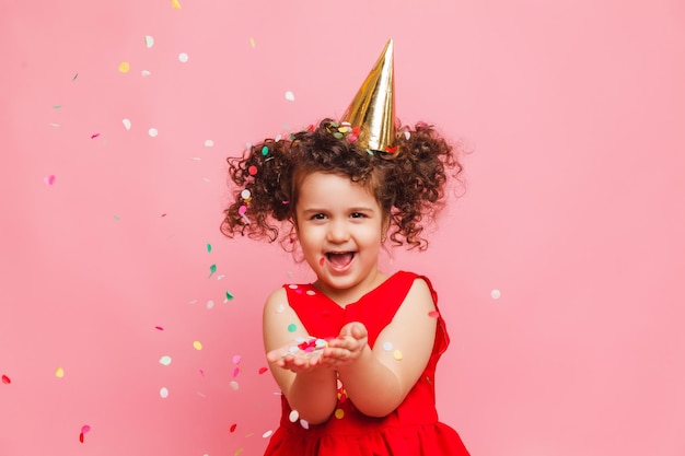 A little girl in a red dress celebrates her birthday blowing and catching confetti on a pink background