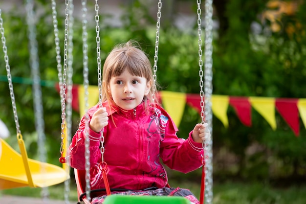 A little girl in a red jacket sits on a chain carousel ride