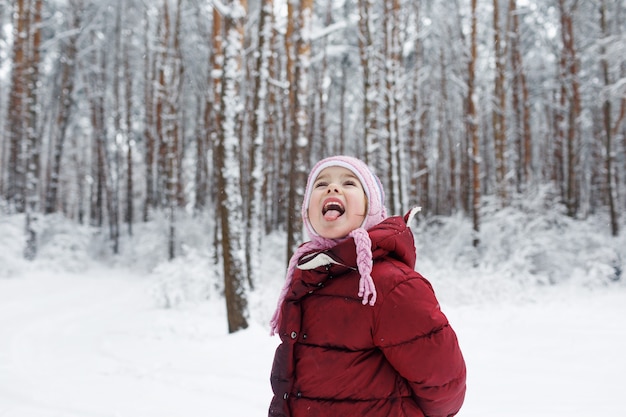 A little girl in a red jacket stands in a snow-covered forest catching snowflakes with her tongue.