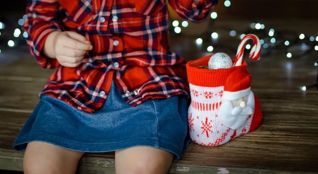 A little girl in a red plaid shirt and a denim blue skirt, opens the sweets from her Christmas gift, sitting on a wooden table. Christmas morning concept. Close up.