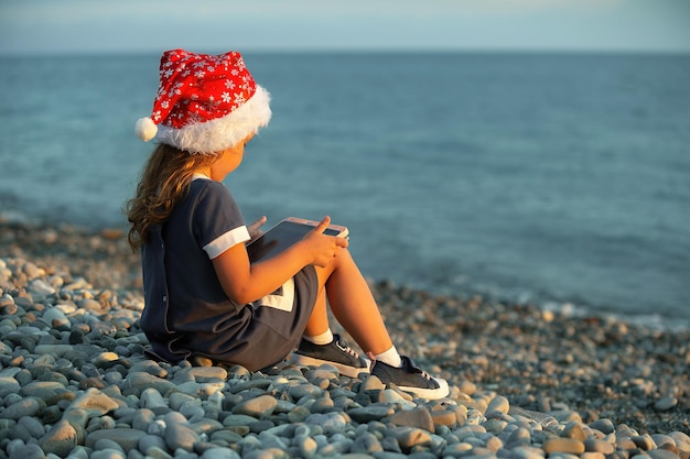 a little girl in a red santa claus hat sits on the seashore with a tablet