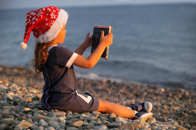 a little girl in a red santa hat sits on the seashore at sunset and watches cartoons on a tablet