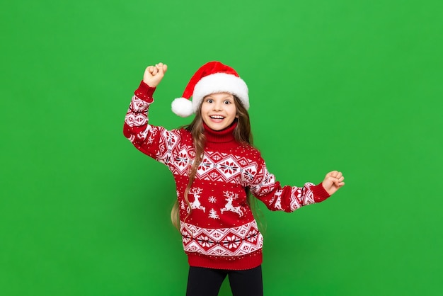 A little girl in a red sweater with reindeer and a Santa Claus hat waiting for the holiday on a green isolated background The concept of Christmas