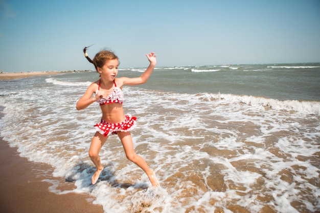 A little girl in a red swimsuit is playing on the beach with a sea wave jumping running having fun Swimming traveling playing with water