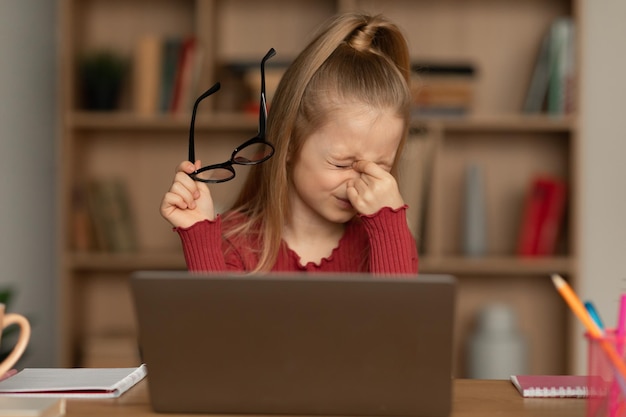 Little girl rubbing eyes holding glasses using laptop at home