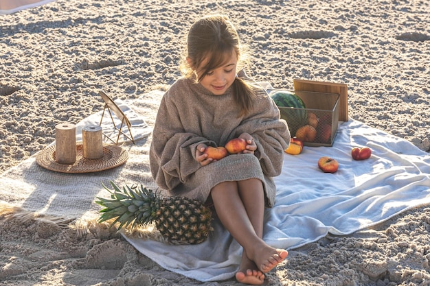 Little girl on the sandy seashore at a picnic with fruits