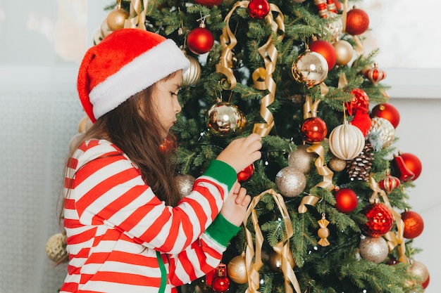 Little girl in santa claus hat decorates the tree with toys. A cute baby is preparing to celebrate Christmas.