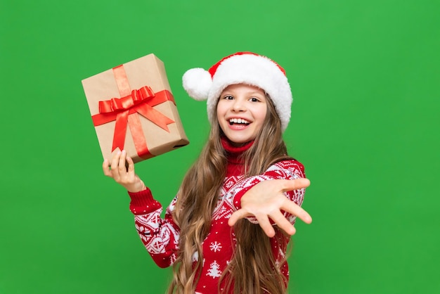 A little girl in a Santa Claus hat and sweater stretches her hand forward and holds a Christmas gift on a green isolated background A New Year's gift to a child