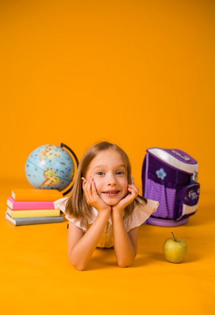 A little girl in a school uniform is lying on the floor with school supplies on a yellow background with a place for text