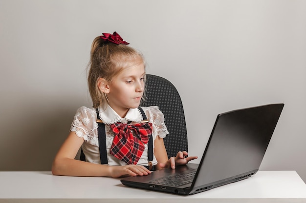 A little girl in a school uniform uses a laptop at a table on a white background