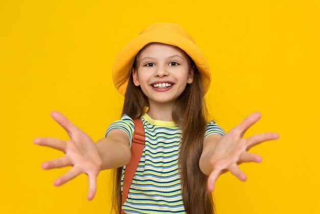 A little girl scout in a hat and with a backpack is going on a summer trip and stretches her palms forward The child goes camping Tourism for children Yellow bright isolated background