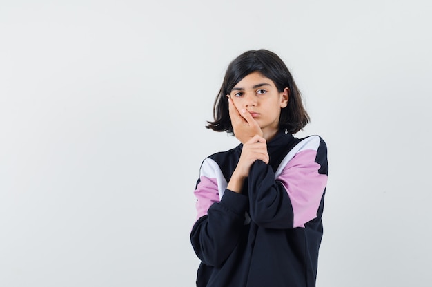 Little girl in shirt holding hand on cheek and looking pensive , front view.