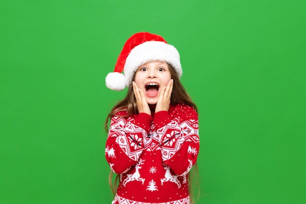 A little girl shows delight at a Christmas gift A child with long hair in a reindeer sweater and Santa Claus hat on a green isolated background