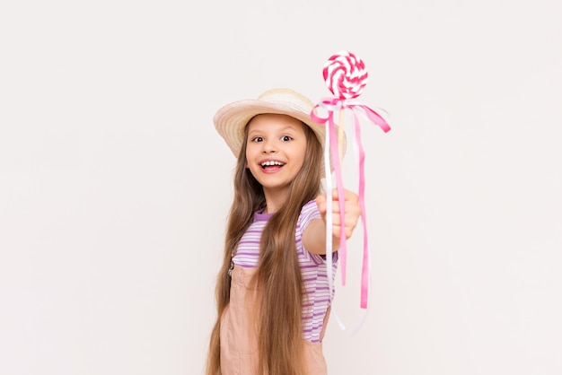 A little girl shows a lollipop and smiles A child in a sundress and a summer hat on a white isolated background