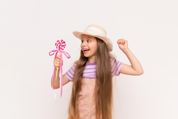 A little girl shows a lollipop and smiles A child in a sundress and a summer hat on a white isolated background