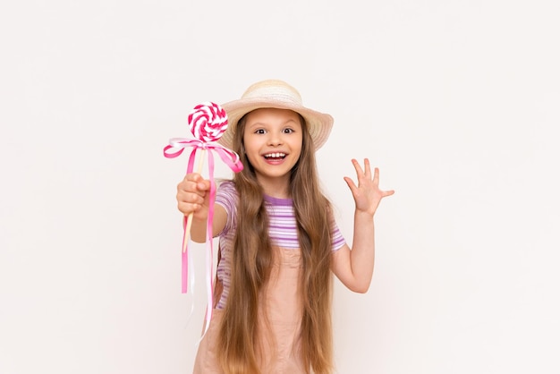 A little girl shows a lollipop and smiles A child in a sundress and a summer hat on a white isolated background