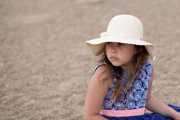 Little girl sit in summer hat and beautiful dress on the beach