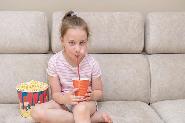 little girl sits on couch at home drinks cocktail from a straw, next to it is huge glass of popcorn