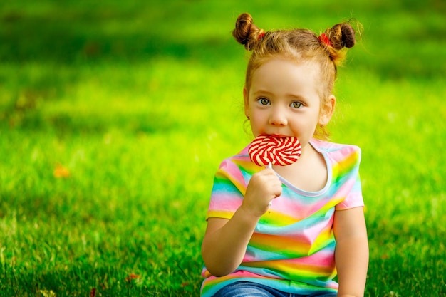 A little girl sits on the grass in the park and puts a lollipop in her mouth