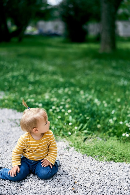 Little girl sits on a gravel path in a green park turning her head to the side