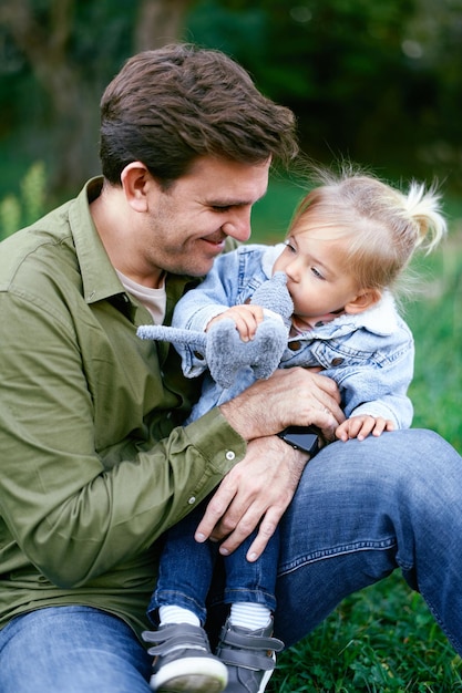 Little girl sits on her dad lap on a green lawn with a toy in her hand