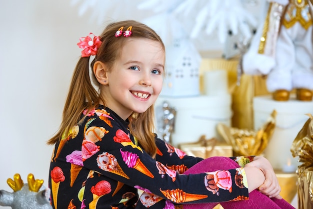 Little girl sits near a New Year tree and smiles. Near the box with a gift.