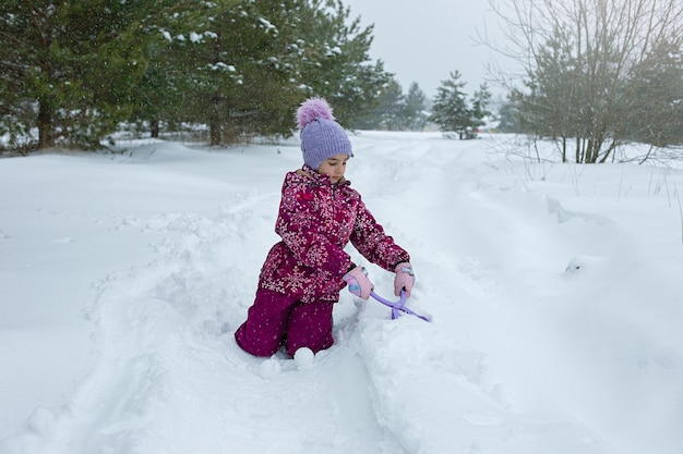 A little girl sits in the snow makes snowballs using a plastic sculpting tool