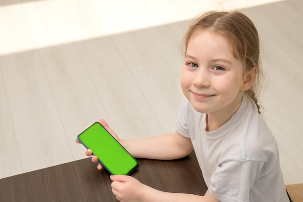 Little girl sits at a table holding a phone with a green screen in her hands top view