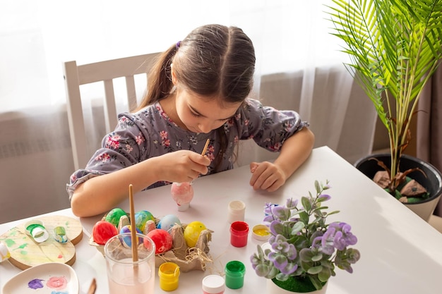 A little girl sits at a white table near the window prepares for the Easter holiday