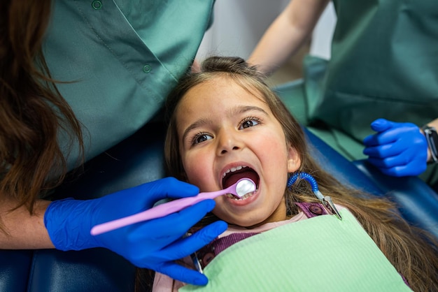 Little girl sitting in a dental chair and two female dentists treating baby teeth