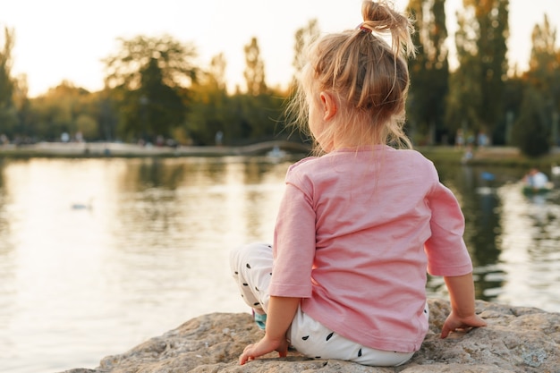 Little girl sitting on a huge stone in park near the lake