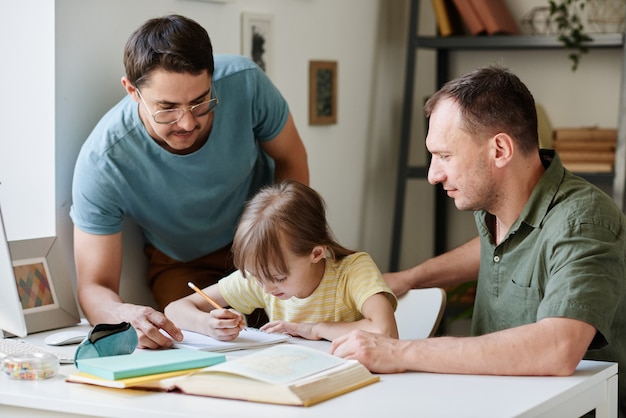 Little girl sitting at the table and doing her homework while gay parents helping her