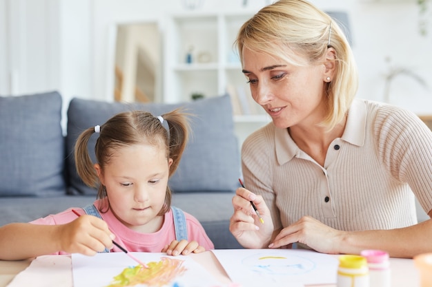 Little girl sitting at the table and painting a picture together with her mother at home