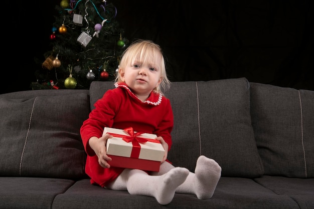 Little girl sitting with a gift in his hands on the sofa against the background of a Christmas tree Christmas eve New Year gifts