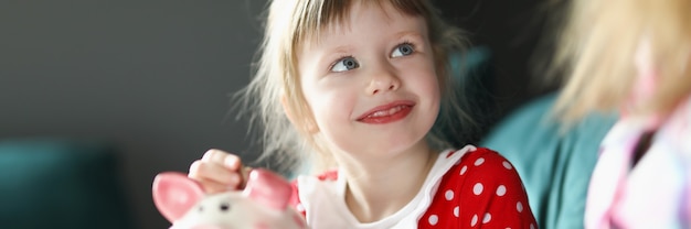 Little girl sitting with her mother on couch and putting coin into piggy bank