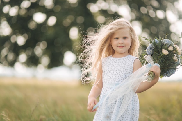 Little girl in sky blue dress with bouquet stand in field in front of big tree Child smile and have fun