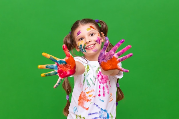 A little girl smeared in multicolored paint pulls her hands forward and smiles on a green isolated background Children's artistic creativity