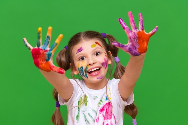 A little girl smeared in multicolored paint pulls her hands forward and smiles on a green isolated background Children's artistic creativity